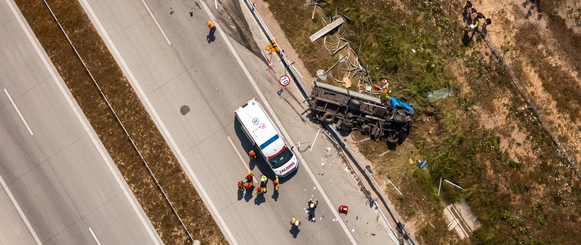 Ujęcie z drona ukazujące pasy jezdni, strażaków, ratowników oraz wywróconą ciężarówkę w rowie