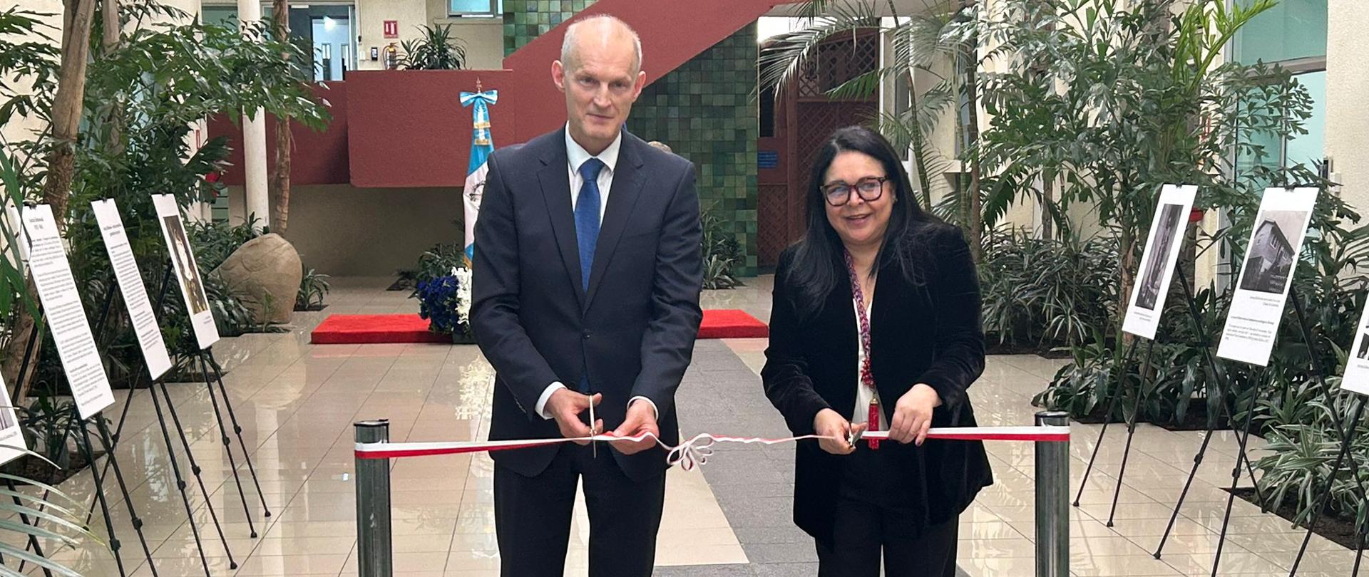 Two people in formal attire cut a red-and-white ribbon during the opening of an exhibition. In the background, a spacious atrium with plants and display boards can be seen on both sides of the walkway.