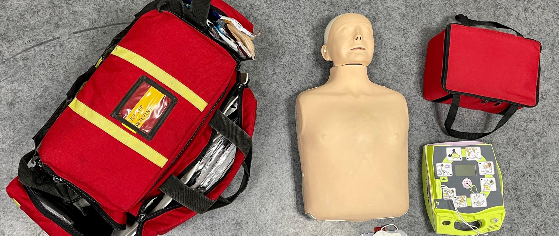 na podłodze widać sprzęt kpp czyli torba r1, manekin do ćwiczeń oraz defiblyrator