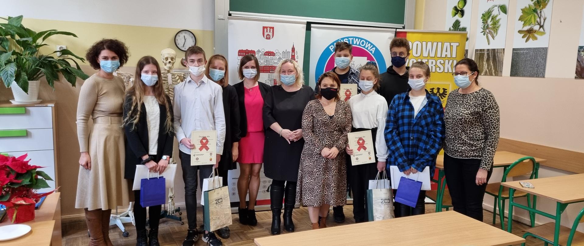 Na zdjęciu znajdują się laureaci Powiatowego Konkursu Wiedzy o AIDS ich opiekunowie i członkowie Jury. Zdjęcie zrobione jest w pracowni biologicznej na tle banerów Urzędu Miasta Zgierza, Powiatowej Stacji Sanitarno-Epidemiologicznej w Zgierzu oraz Powiatu Zgierskiego.