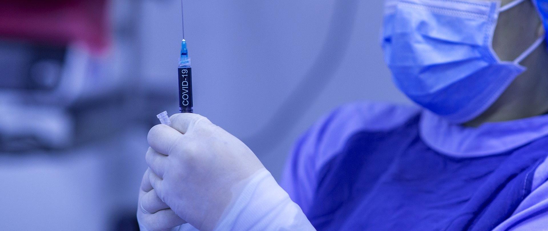 Na zdjęciu pielęgniarka, ubrana w niebieski kitel. Trzyma w dłoni strzykawkę z napisem Covid-19. Na dłoniach ma założone białe,lateksowe rękawice ochronne. Granatowe tło. 