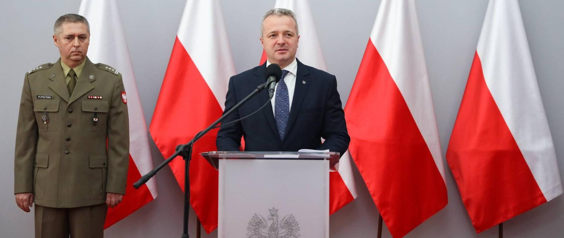 Jeden mężczyzna stojący przy mównicy i wypowiadający się. Obok stoi drugi mężczyzna