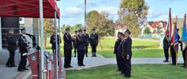 Trzech strażaków ubranych mundury galowe stoi w rzędzie po odebraniu odznaczeń, po lewej stronie widoczny namiot z mównicą dla zaproszonych gości po prawej stronie stoi poczet sztandarowy 