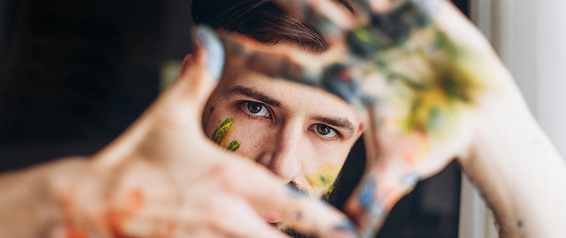 Młody człowiek w białej, ubrudzonej kolorowymi farbami podkoszulce, wyciąga w stronę obiektywu ubrudzone kolorowymi farbami dłonie, z których układa się prostokątny kadr, w którym widać jego oczy oraz fragment, także ubrudzonej kolorowymi farbami twarzy