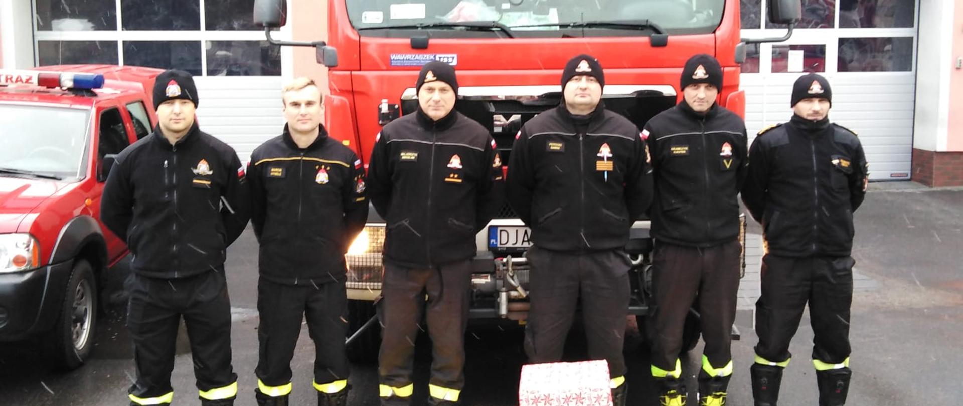 Obraz przedstawia strażaków stojących na tle pojazdów pożarniczych i budynku jaworskiej komendy. Przed strażakami ustawione świąteczne paczki.