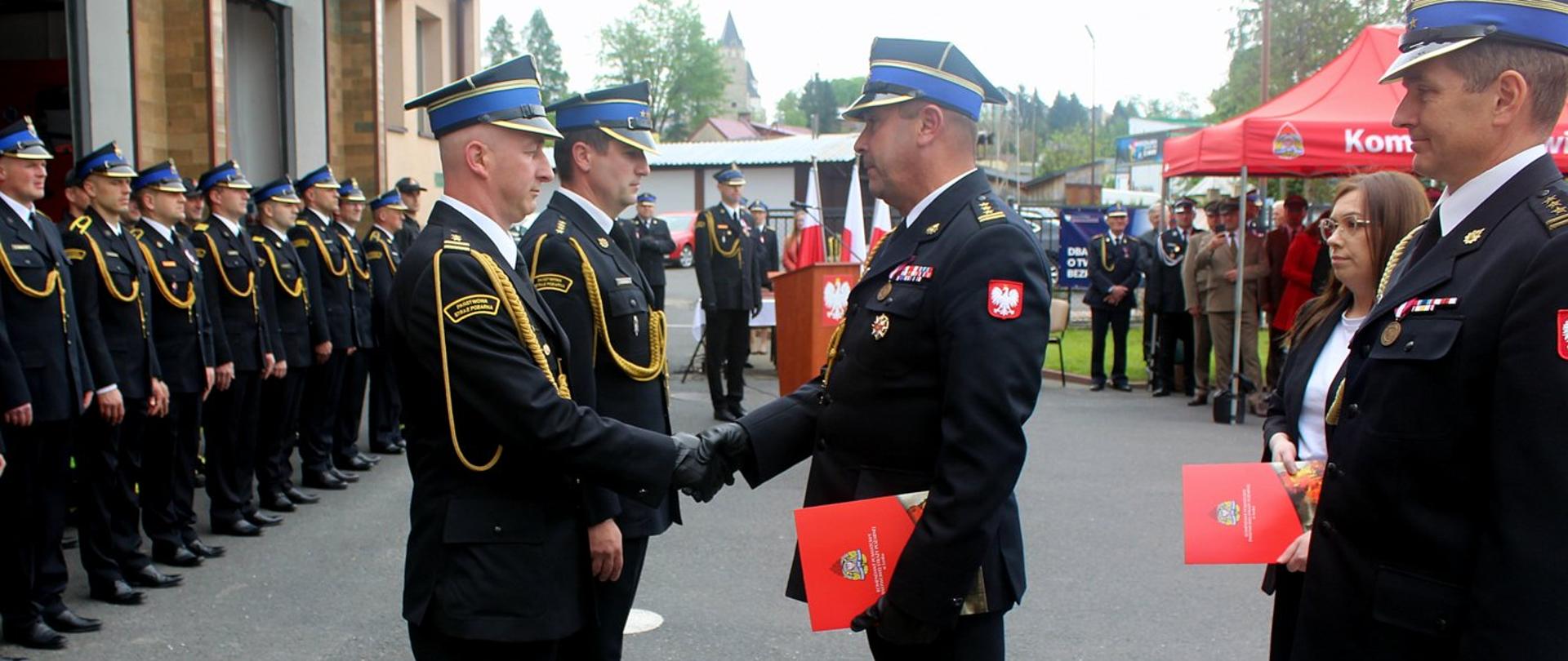 Zdjęcie zrobione na zewnątrz przed komendą powiatową Państwowej Straży Pożarnej, podczas uroczystości. Na placu przed jednostką stoją uczestnicy uroczystości - funkcjonariusze Państwowej Straży Pożarnej. Funkcjonariusz Państwowej Straży Pożarnej w stopniu starszego brygadiera w lewej ręce trzyma teczkę z dokumentami, prawą podaje strażakowi w stopniu brygadiera, gratulując wyróżnienia. 