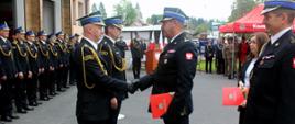 Zdjęcie zrobione na zewnątrz przed komendą powiatową Państwowej Straży Pożarnej, podczas uroczystości. Na placu przed jednostką stoją uczestnicy uroczystości - funkcjonariusze Państwowej Straży Pożarnej. Funkcjonariusz Państwowej Straży Pożarnej w stopniu starszego brygadiera w lewej ręce trzyma teczkę z dokumentami, prawą podaje strażakowi w stopniu brygadiera, gratulując wyróżnienia. 