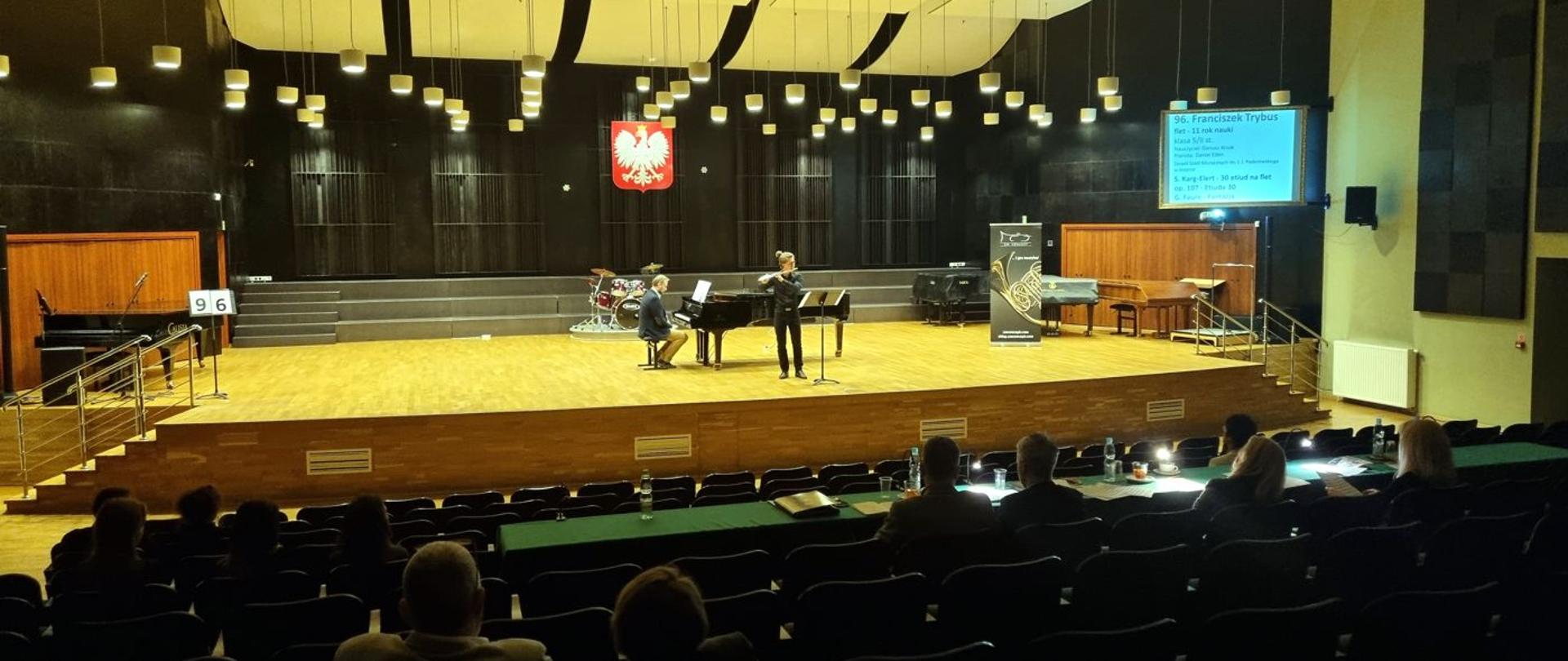 Postaci flecisty Franciszka Trybusa i pianisty Daniela Eibina na środku dużej sceny. Na pierwszym planie komisja konkursowa siedząca na widowni