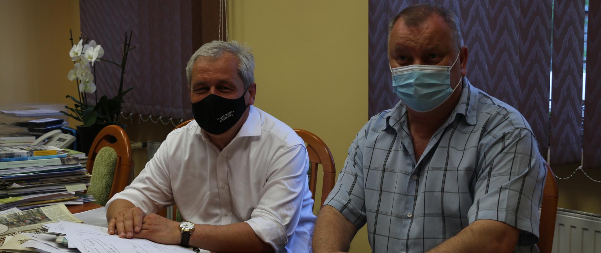 Spotkanie wojewody opolskiego z wójtem gminy Lubsza. Na zdjęciu obaj panowie, zasiadający za dużym stołem, na którym jest sporo dokumentów. Wojewoda ubrany w koszulę białą, z podwiniętymi rękawami. Na twarzy ma maseczkę ochronną. Obok niego - po jego lewej stronie, siedzi za stołem wójt gminy, ubrany w szarą koszulę w kratkę. Na twarzy ma również maseczkę ochronną, ręce oparte o stół. Poniżej kadru widoczny kalkulator. W tle, za mężczyznami widać okno, zasłoniętę roletami. Obok wojewody po jego prawej stronie - na dalszym planie na biurku, widać brązową doniczkę z białym kwiatek orchidei.