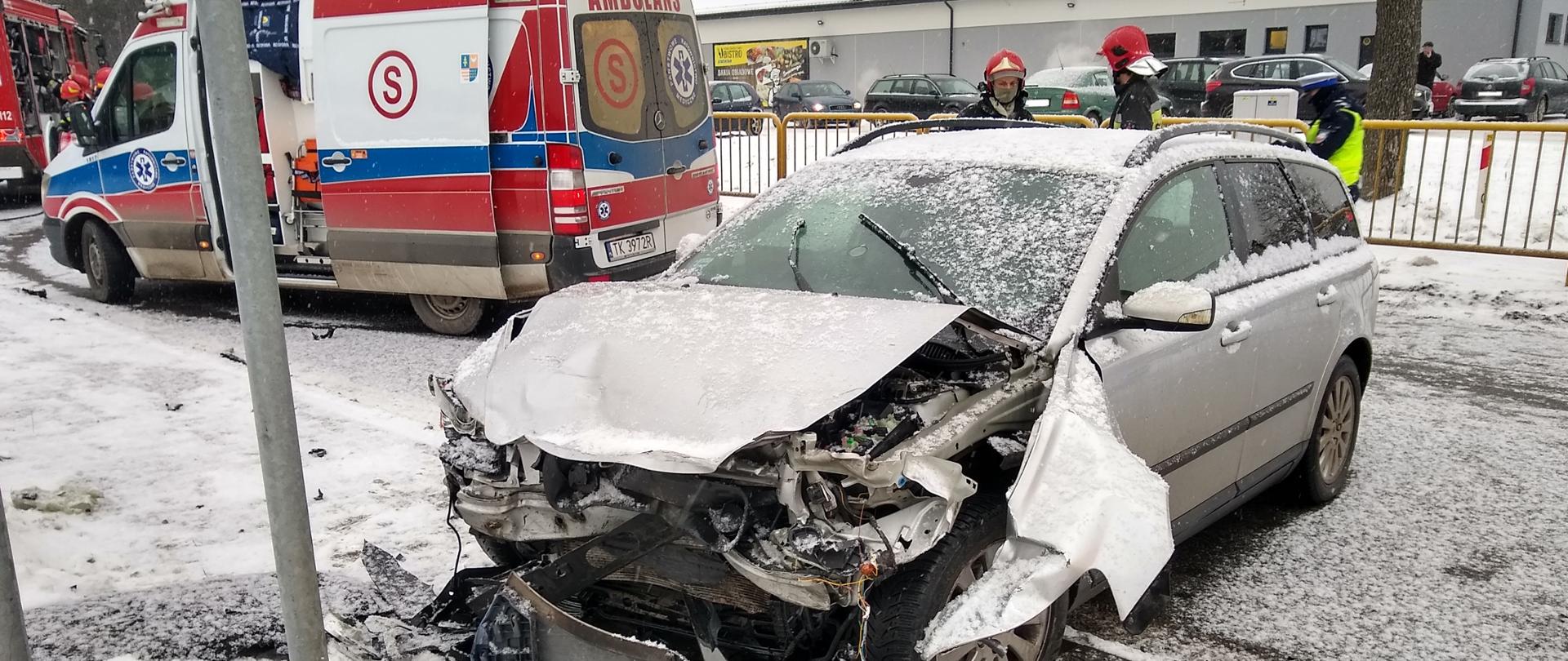 Wypadek drogowy. Na pierwszym planie uszkodzony (rozbity z przodu) samochód osobowy marki Volvo V50. Strażacy i policjanci zabezpieczają teren działań. Na miejscu obecny jest Zespół Ratownictwa Medycznego