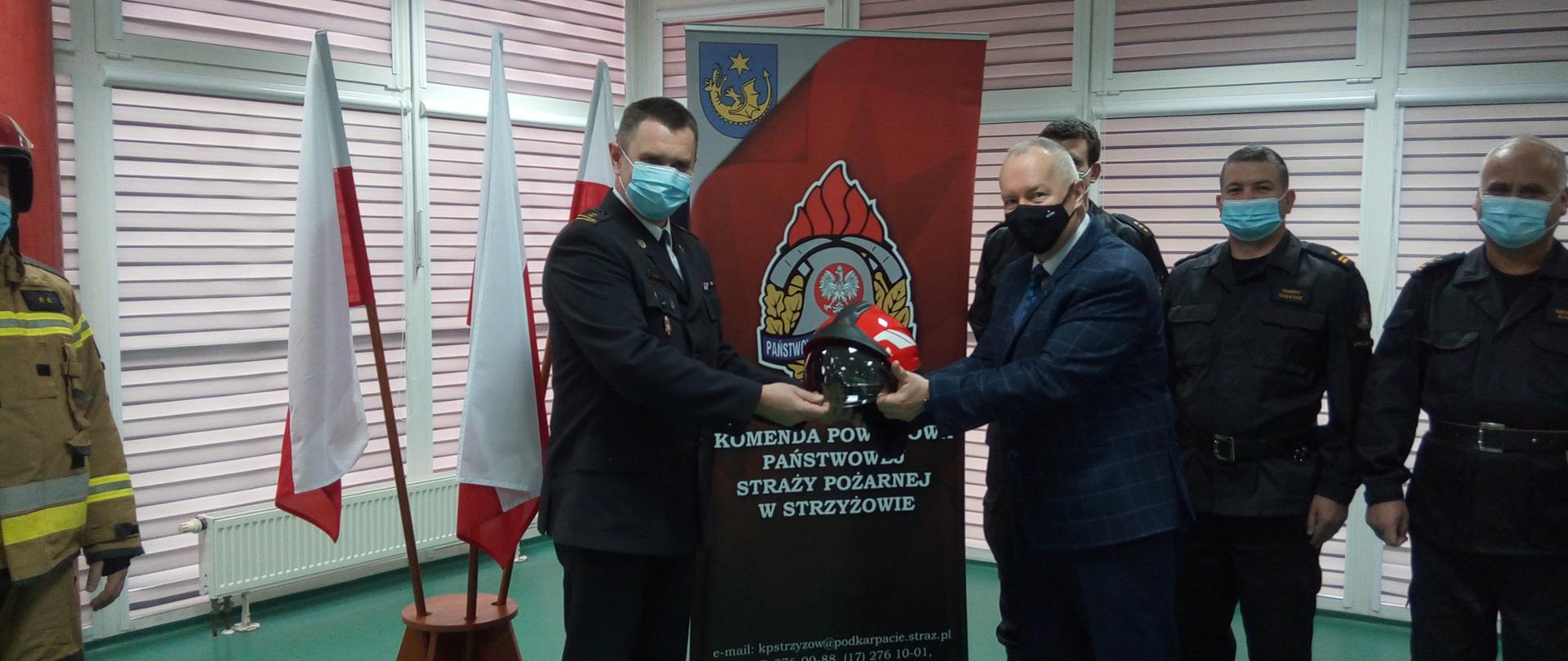 Na zdjęciu w centralnej jego części Komendant Powiatowy Państwowej Straży Pożarnej w Strzyżowie brygadier Rafał Rutyna odbiera hełm strażacki z rąk Starosty strzyżowskiego Pana Bogdana Żyburę. Obaj Panowie trzymają w hełm strażacki w geście przekazania. W tle od lewej strony trzy Flagi Polski, baner Komendy Powiatowej Państwowej Straży Pożarnej w Strzyżowie oraz trzech funkcjonariuszy w umundurowaniu dowódczo-sztabowym. Wszyscy na zdjęciu mają zasłoniętą twarz maseczką.