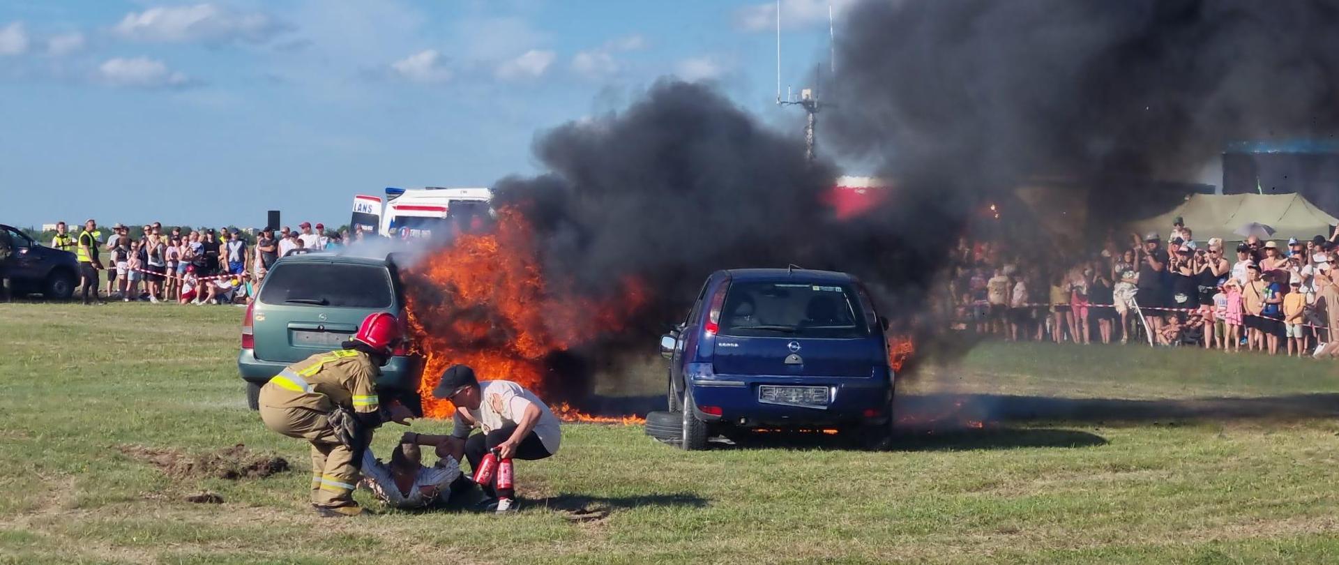 zdjęcie z pokazów, widać strażaka i pozoranta ciągnącego w bezpieczne miejsce osobę poszkodowaną, w tle palące się 2 samochody osobowe