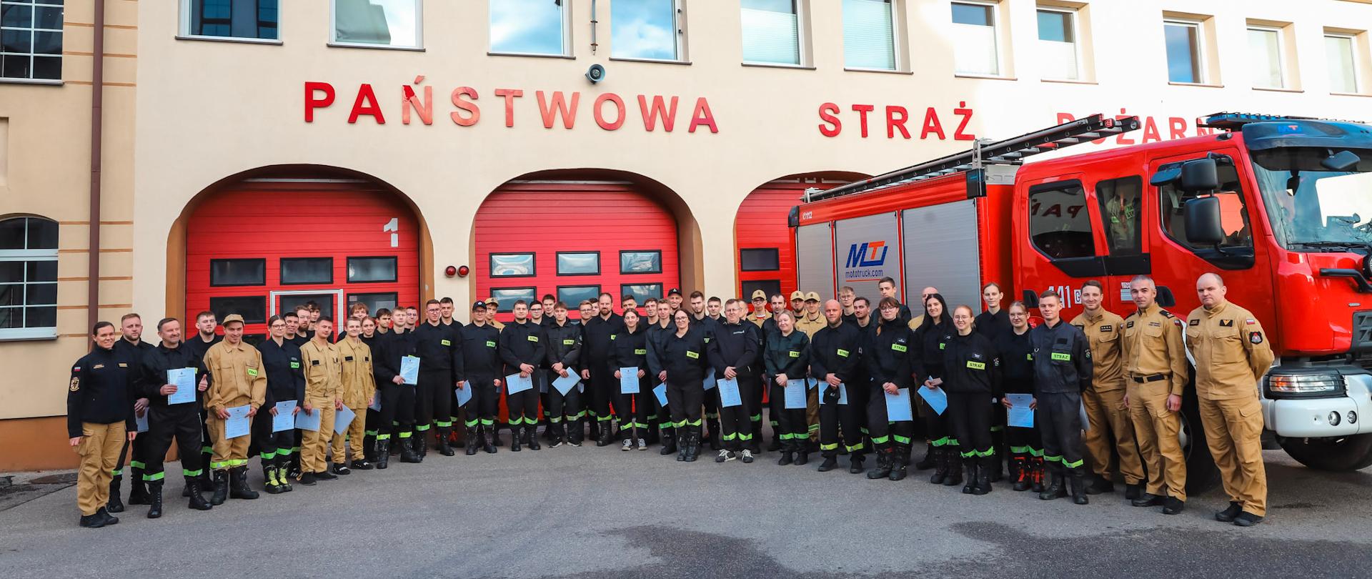 Jest to zdjęcie grupowe po zakończonym szkoleniu, znajdują się na nim wszyscy słuchacze, Zastępca Komendanta Powiatowego oraz funkcjonariusze wydziału planowania operacyjnego. W tle samochód strażacki oraz budynek komendy. 