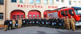Jest to zdjęcie grupowe po zakończonym szkoleniu, znajdują się na nim wszyscy słuchacze, Zastępca Komendanta Powiatowego oraz funkcjonariusze wydziału planowania operacyjnego. W tle samochód strażacki oraz budynek komendy. 