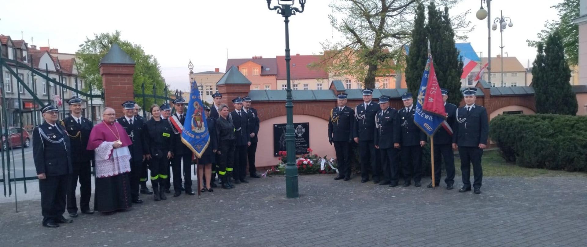 Zdjęcie przedstawia strażaków podczas uroczystości z okazji Międzynarodowego Dnia Strażaka.
W tle ogrodzenie i budynki.
