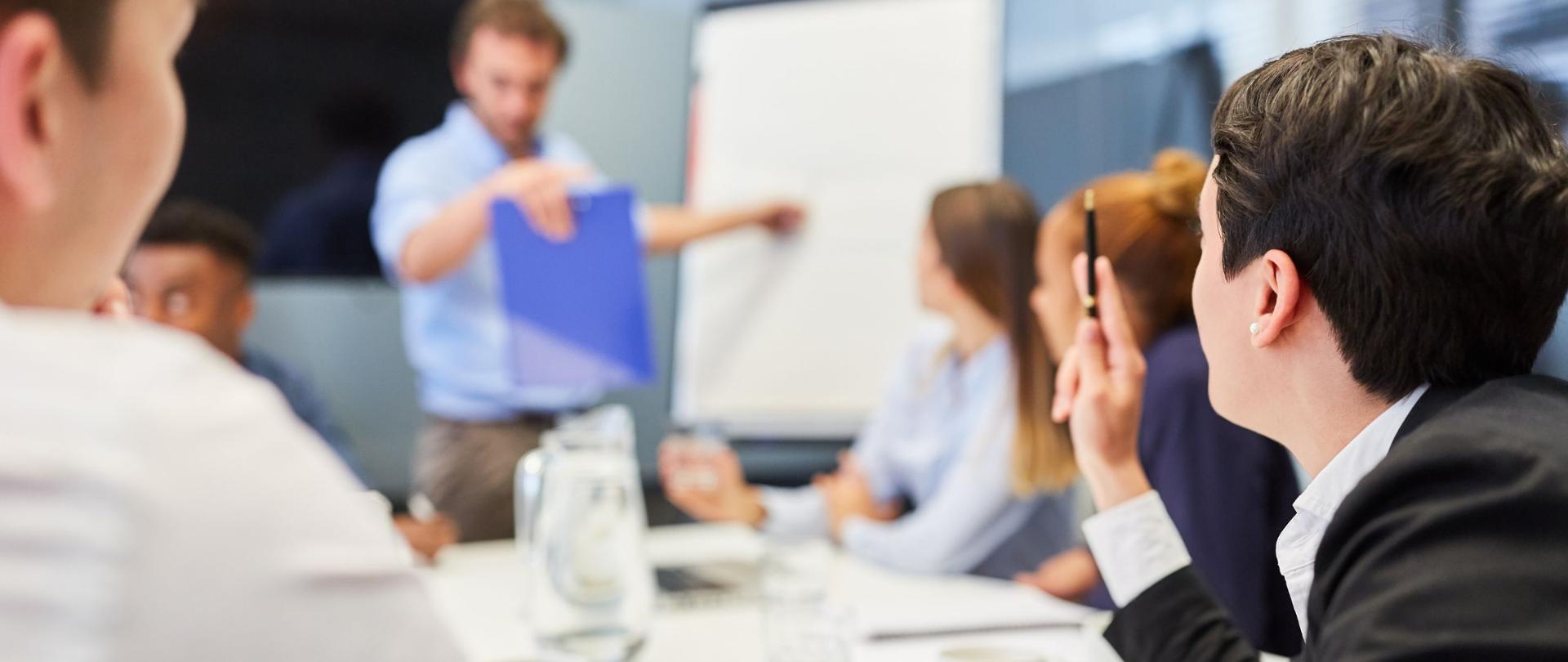 zdjęcie poglądowe - osoby siedzące przy stole słuchają prowadzącego