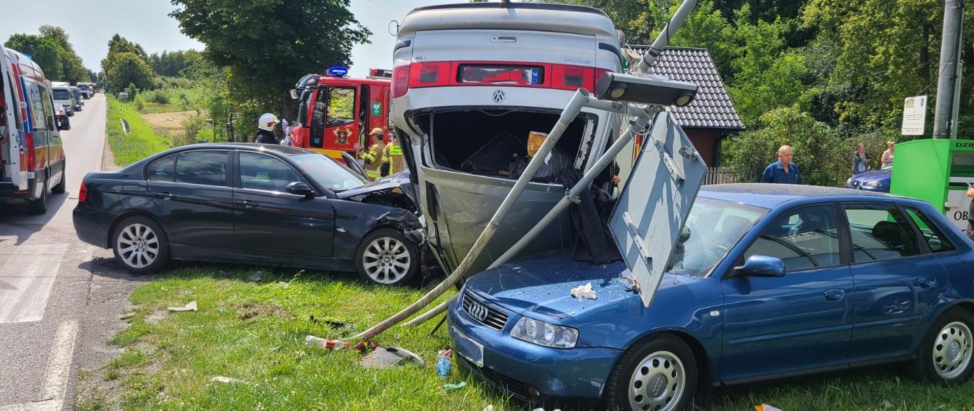 Zdjęcie przedstawia wszystkie trzy samochody biorące udział w zderzeniu: od lewej soi BMW, dalej leżący na dachu Volkswagen i z prawej strony zniszczone Audi. W centralnym punkcie widać zniszczone znaki drogowe. 