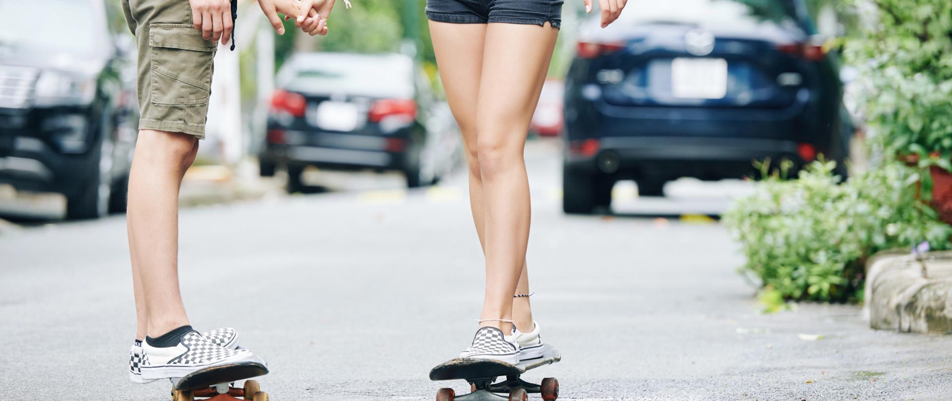 Młody chłopak i dziewczyna na deskorolkach, trzymający się za ręce. Widok z dołu - głównie nogi i tors, nie widać głów i ramion