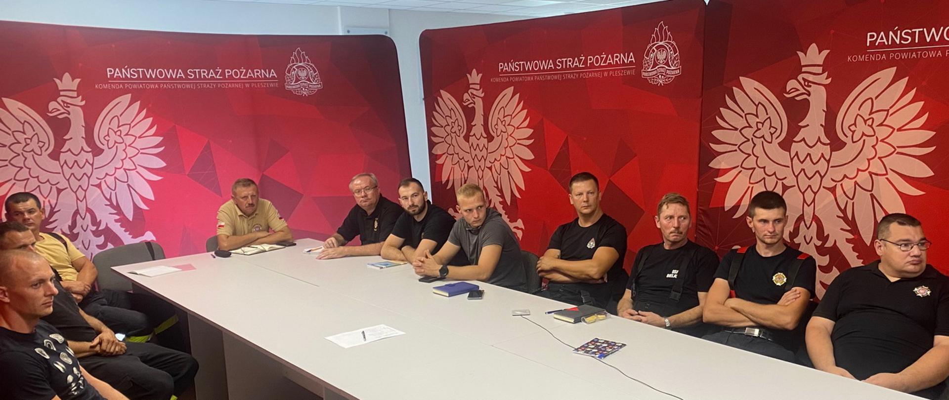 Grupa mężczyzn siedzi przy białym stole, w tle znajdują się banery straży pożarnej, na których widoczny jest orzeł państwowy oraz logo PSP i napis Państwowa Straż Pożarna na czerwonym tle.