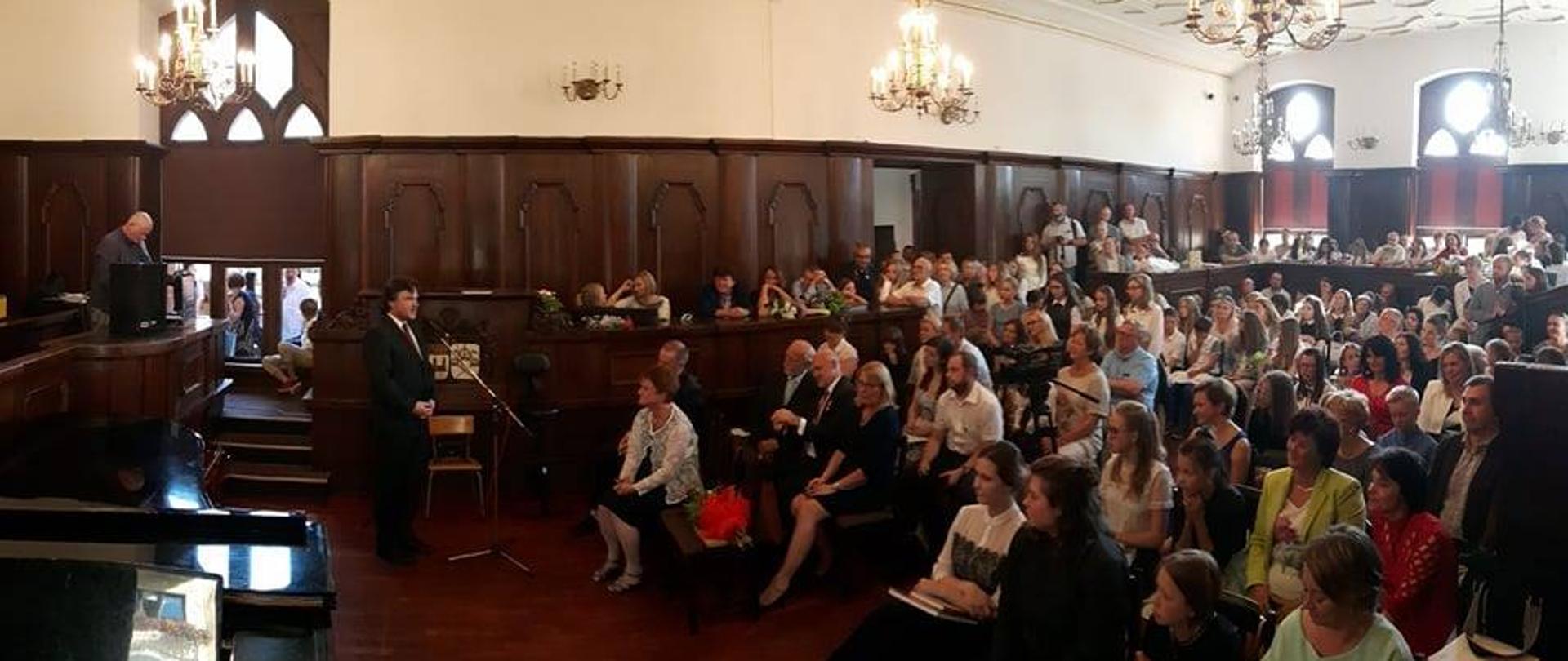Zdjęcie uroczystości zakończenie roku szkolnego w sali Ratusza Miejskiego. Ściany ozdobione zabytkową dębową boazerią, sufit biały, zdobiony kasetonami ze złoconymi ornamentami i ozdobnymi żyrandolami. Po lewej stronie przemawiający do mikrofonu dyrektor, po prawej stronie zgromadzona publiczność. W lewym dolnym narożniku zdjęcia widoczny otwarty fortepian.