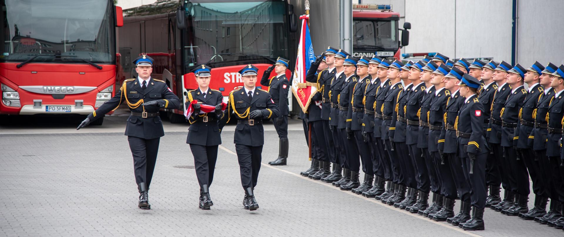 Strażacy w mundurach galowych maszerują przed kompanią honorową ustawioną w szeregu. W tle stoją czerwone pojazdy strażackie zaparkowane przy garażach.
