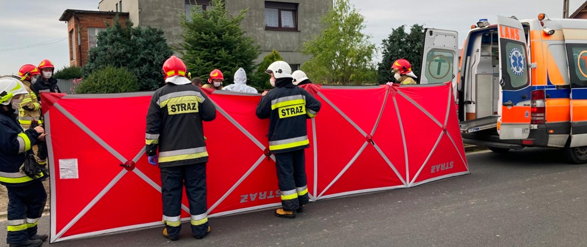 Na zdjęciu widać parawan, karetkę pogotowia oraz strażaków udzielających pomocy osobie nieprzytomnej.