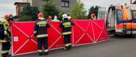 Na zdjęciu jest parawan oraz karetka pogotowia. Strażacy udzielają pomocy osobie poszkodowanej.