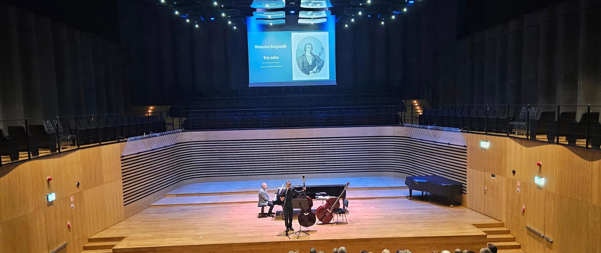 Mężczyzna trzyma kontrabas i mówi do mikrofonu, za nim przy fortepianie siedzi drugi mężczyzna, z przodu widać od tyłu publiczność.