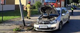 Na zdjęciu widzimy srebrny samochód stojący przodem przy słupie oświetleniowym, w który uderzył. Na miejscu są strażacy oraz Policja, a w tle stoi czerwony wóz strażacki. Auto jest wyraźnie uszkodzone od strony kierowcy, a maska podniesiona. Na jezdni i chodniku nie ma innych pojazdów. W tle znajdują się budynki mieszkalne i niebieskie niebo bez chmur.