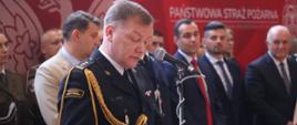 Na zdjęciu widać st. bryg. Pawła Kielara, nowego Opolskiego Komendanta Wojewódzkiej Państwowej Straży Pożarnej w Opolu. Stoi przy mównicy i mówi do mikrofonu. Za nim widać uczestników uroczystości. 