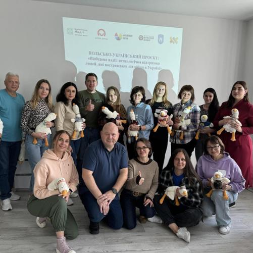 Grupa kobiet i mężczyzn pozuje do zdjęcia na tle slajdu, na którym wyświetlone jest logo Polskiej pomocy