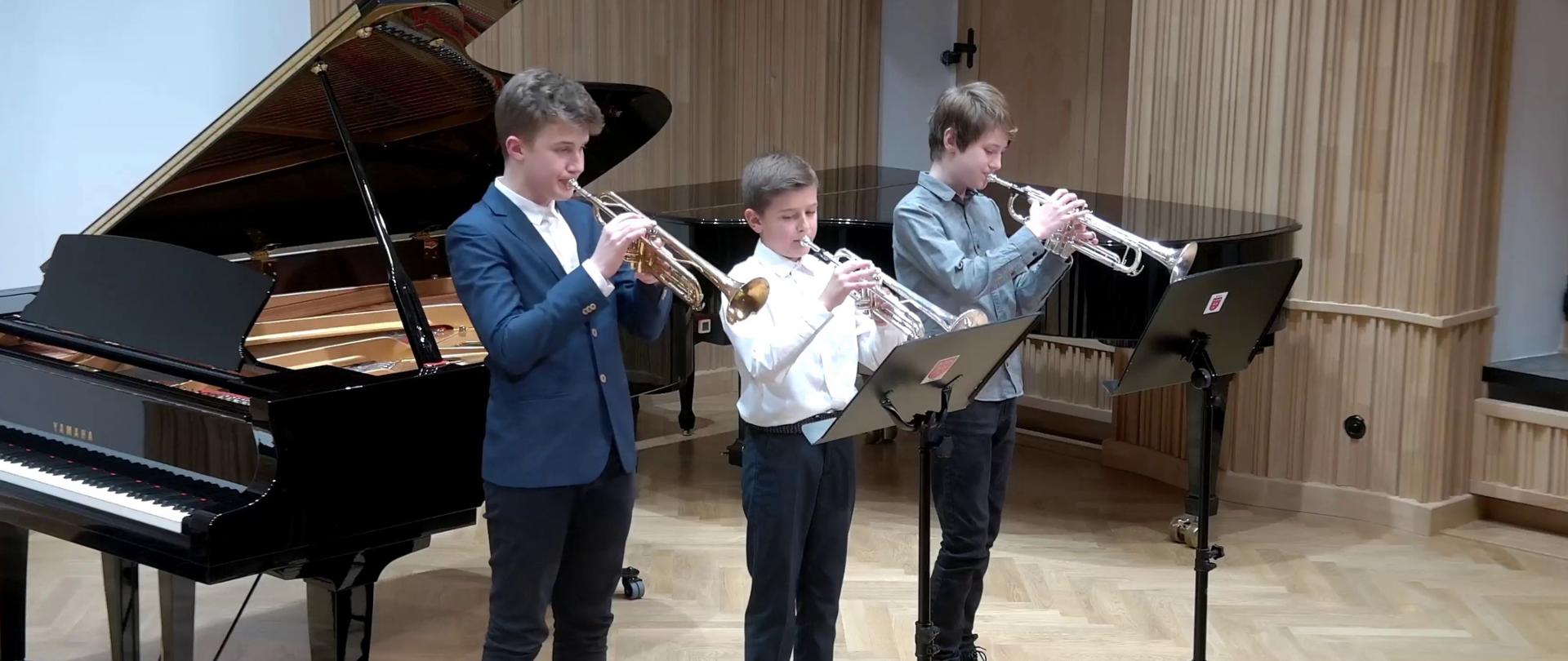 Zdjęcie. Sala Koncertowa Na estradzie występuje trzech młodych trębaczy. Przed nimi stoją 2 czarne pulpity, za nimi czarny fortepian otwarty, w głębi czarny fortepian zamknięty.