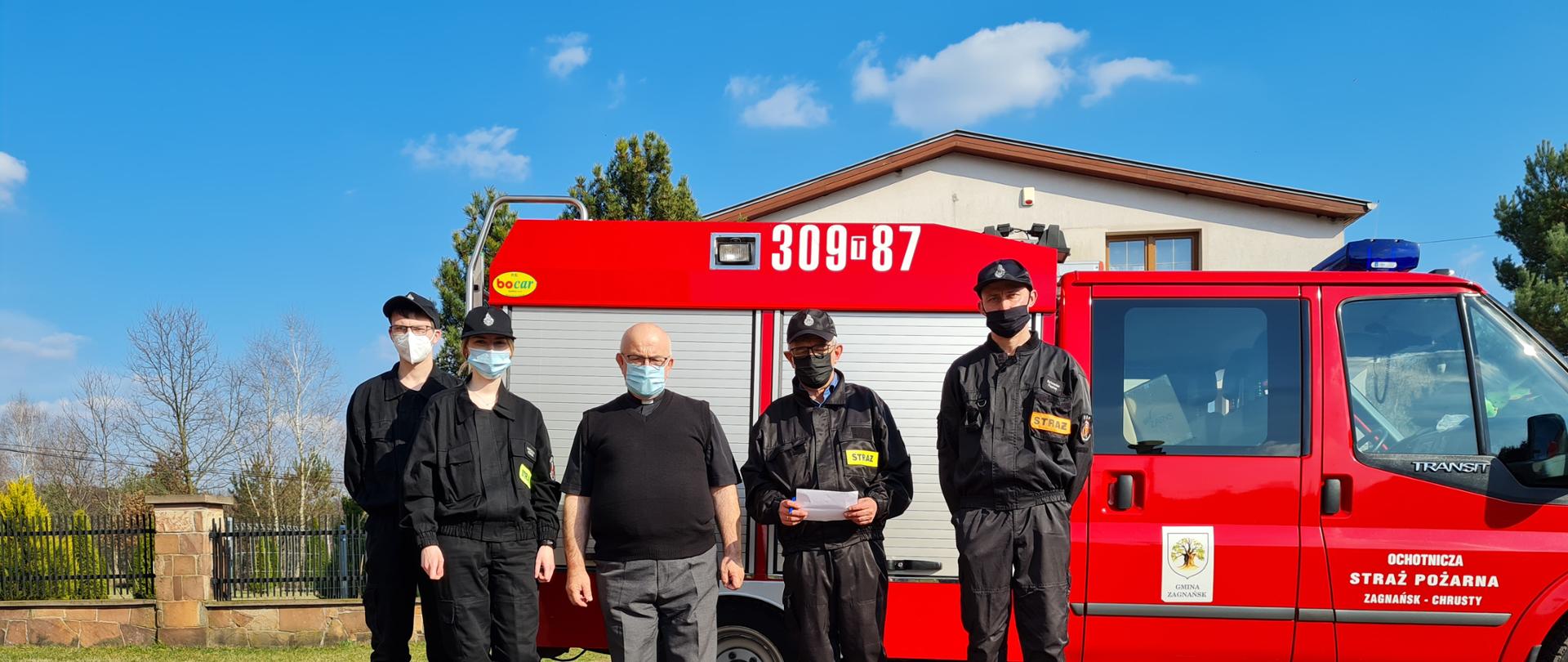 Zdjęcie przedstawia strażaków stojących przed samochodem gaśniczym. Przed nimi znajdują się płyny do dezynfekcji i maseczki.