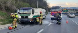 Strażacy prowadzą działania przy kabinie samochodu ciężarowego stojącego na drodze. Jeden ze strażaków zdejmuje aparat ochrony dróg oddechowych. Obok stoi samochód pożarniczy. Drugim pasem jedzie samochód.