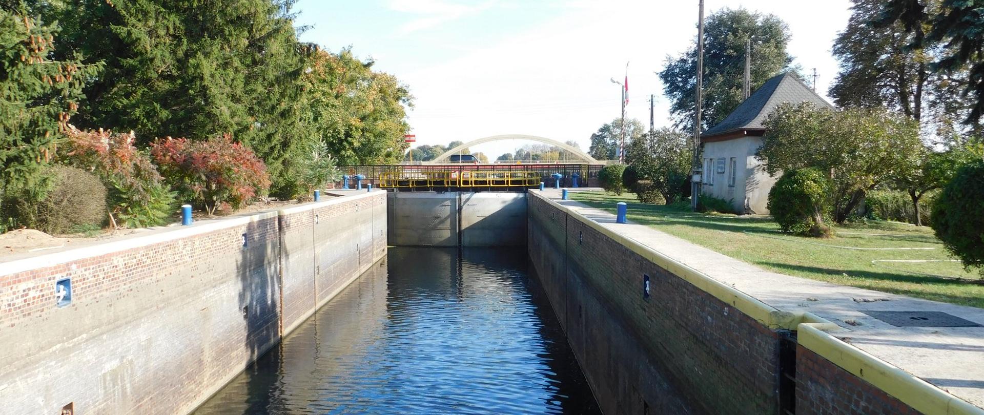 Śluza nr 6 (Osowa Góra) drogi wodnej Wisła - Odra, będąca elementem międzynarodowej drogi wodnej E-70. Wrota śluzy zamknięte. Komora śluzy napełniona wodą. Słoneczny, bezchmurny dzień. Na drugim planie most nad rzeką Notecią. Po prawej stronie śluzantówka, po lewej drzewa i krzewy. 