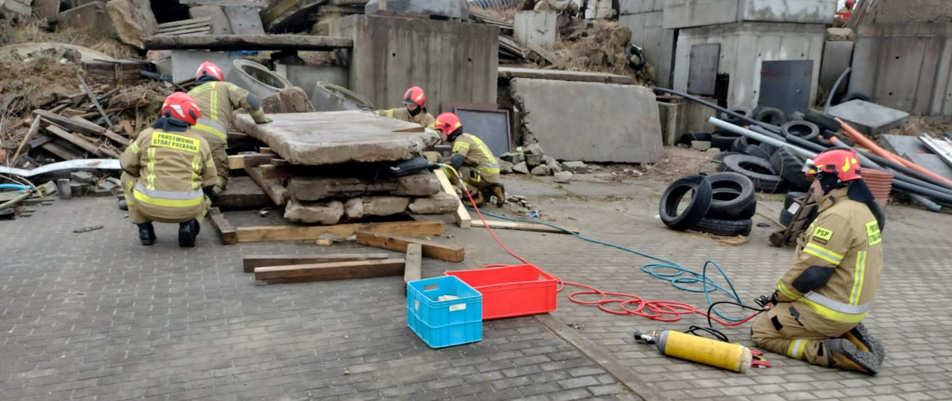 Strażacy w umundurowaniu specjalnym ćwiczą ze sprzętem na poligonie.