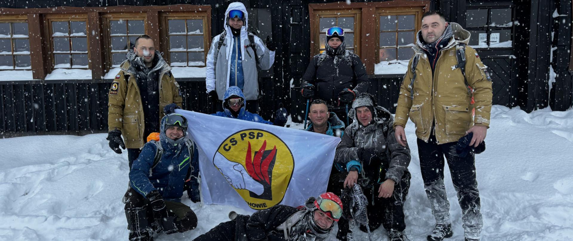 Zimowa sceneria, grupa dziewięciu osób z rozwinięta flagą z logo CS PSP, na pamiątkowym zdjęciu na tle schroniska "Samotnia"