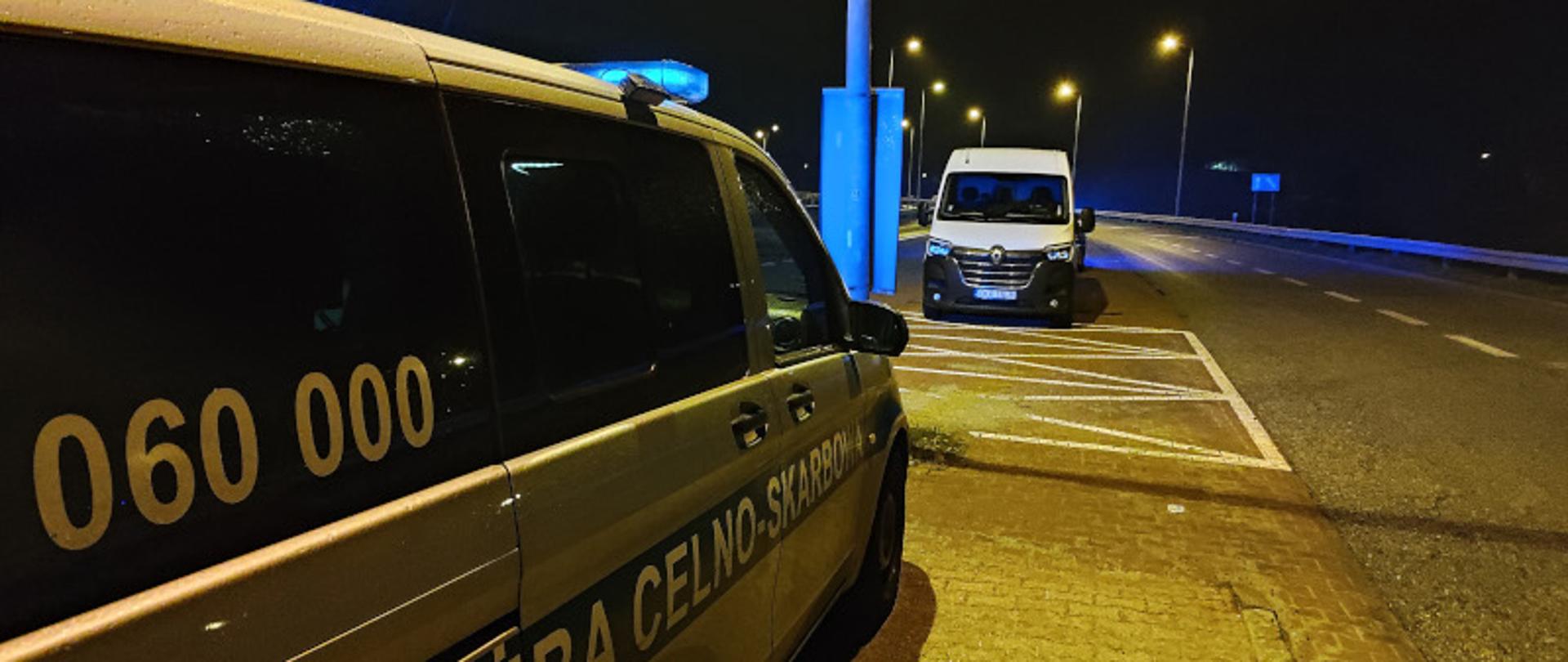 Zdjęcie nocne przedstawiające furgonetkę polskiej Służby Celno-Skarbowej z włączonymi niebieskimi sygnałami świetlnymi, zaparkowaną na poboczu drogi obok białego vana podczas kontroli drogowej.