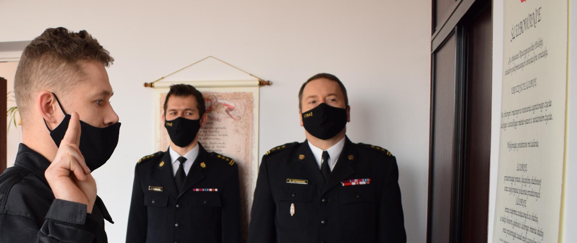 Po lewej stronie stoi mężczyzna, zwrócony prawym profilem do fotografa, a bezpośrednio zwrócony w stronę ściany, na której wisi tekst ślubowania. Mężczyzna ma podniesioną prawą dłoń na wysokość swoich uszu, z wyprostowanymi dwoma palcami w celu ślubowania. Ubrany jest w czarny strój koszarowy oraz ma czarną maseczkę na twarzy. Na drugim planie stoi dwóch mężczyzn na tle białej ściany, zwróconych przodem do fotografa. Obaj mężczyźni ubrani są w mundur strażacki wyjściowy/galowy i mają na twarzy czarne maseczki.