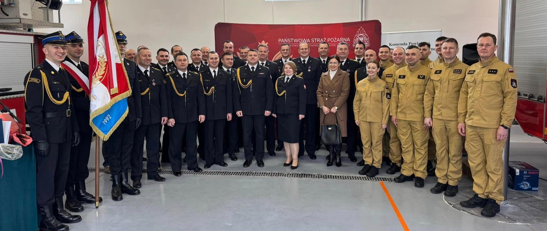 Zdjęcie przedstawia dużą grupę strażaków Państwowej Straży Pożarnej, w tym kilku z nich w galowych mundurach, zebranych wewnątrz budynku, prawdopodobnie w remizie strażackiej. Na pierwszym planie po lewej stronie widać strażaka trzymającego sztandar z numerem "1992". W tle znajduje się czerwony baner z napisem "PAŃSTWOWA STRAŻ POŻARNA" oraz symbolami PSP. Na ścianie widać również numery alarmowe "998-112". W grupie znajduje się także jedna kobieta w jasnym płaszczu.