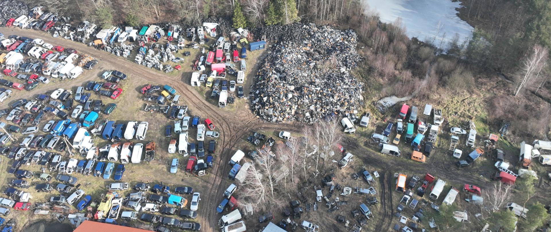 Widok na część terenu ze zgromadzonymi oponami, hałdą odpadów oraz pojazdami w różnym stopniu demontażu