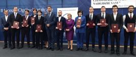 Wiceminister Piontkowski, kobieta ubrana na fioletowo i kilkunastu młodych ludzi w garniturach trzymających dyplomy w brązowych okładkach stoją w rzędzie pod granatową ścianą