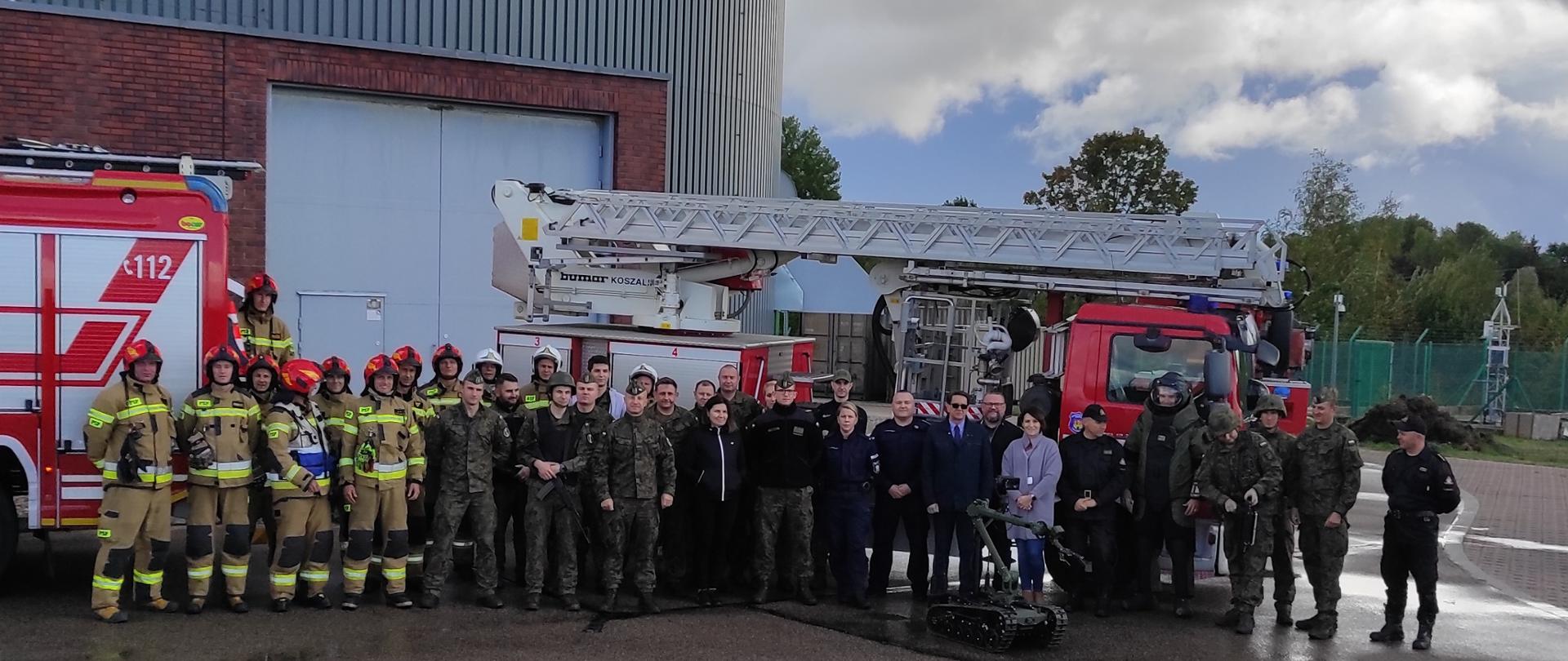 na tle samochodów strażackich zdjęcie grupy osób ustawionych w rzędzie, które brały udział i obserwowały ćwiczenia, strażacy żołnierze i cywile.