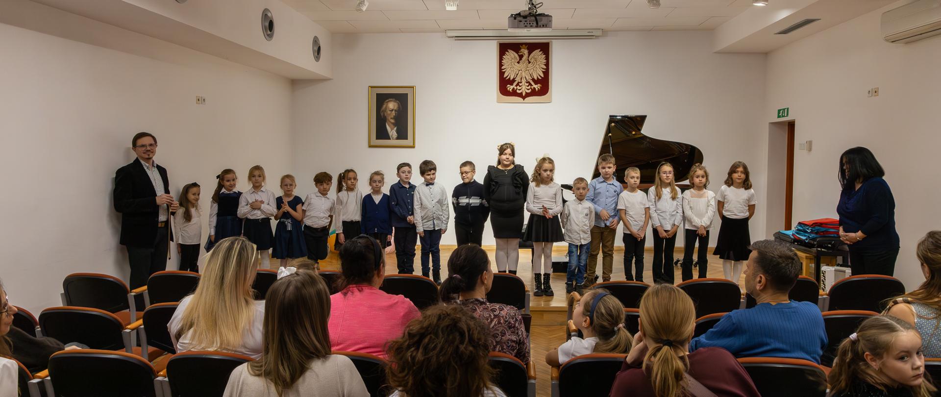 uczniowie klasy pierwszej wspólnie stojący na scenie w auli szkoły muzycznej w głubczycach, z lewej strony zdjęcia widać pana dyrektora szkoły, który pozuje wraz z uczniami, z prawej strony stoi pani przygotowująca uczniów do uroczystości pasowania, w dolnej części zdjęcia widać publiczność zgromadzoną na wydarzeniu