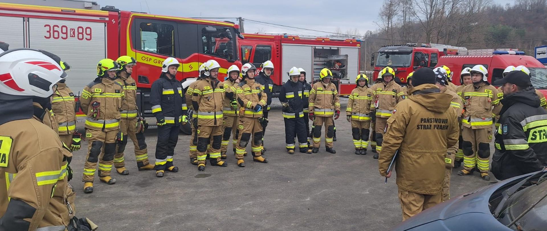 W półkolu stoją strażacy ubrani w piaskowe ubrania bojowe przed nimi stoją organizatorzy ćwiczenia również w ubraniach bojowych. Z tylu widoczne budynki oraz samochody pożarnicze