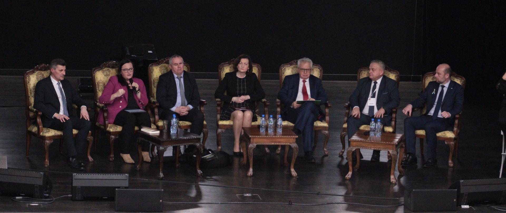 Siedem osób siedzi na fotelach na scenie. Druga od lewej siedzi wiceminister Małgorzata Jarosińska-Jedynak i w lewej dłoni trzyma mikrofon.