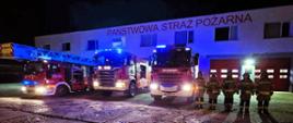 Widoczne trzy samochody straży pożarnej przed jednostką oraz pięciu strażaków.