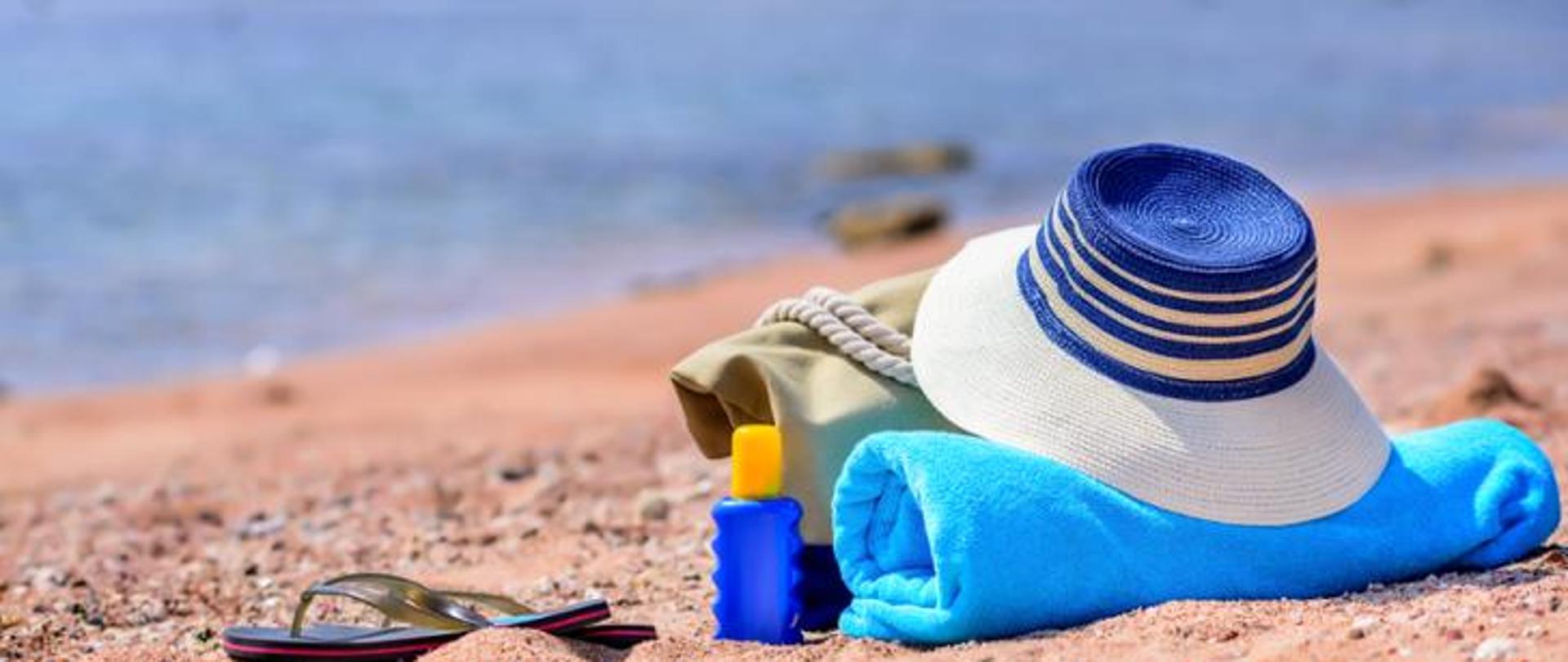 Plaża nad wodą: na piasku leżą czarne klapki, niebieska butelka kremu z filtrem, beżowa torba z liną, słomkowy kapelusz w biało-niebieskie paski oraz zwinięty niebieski ręcznik. W tle widać spokojną wodę i kamienie.