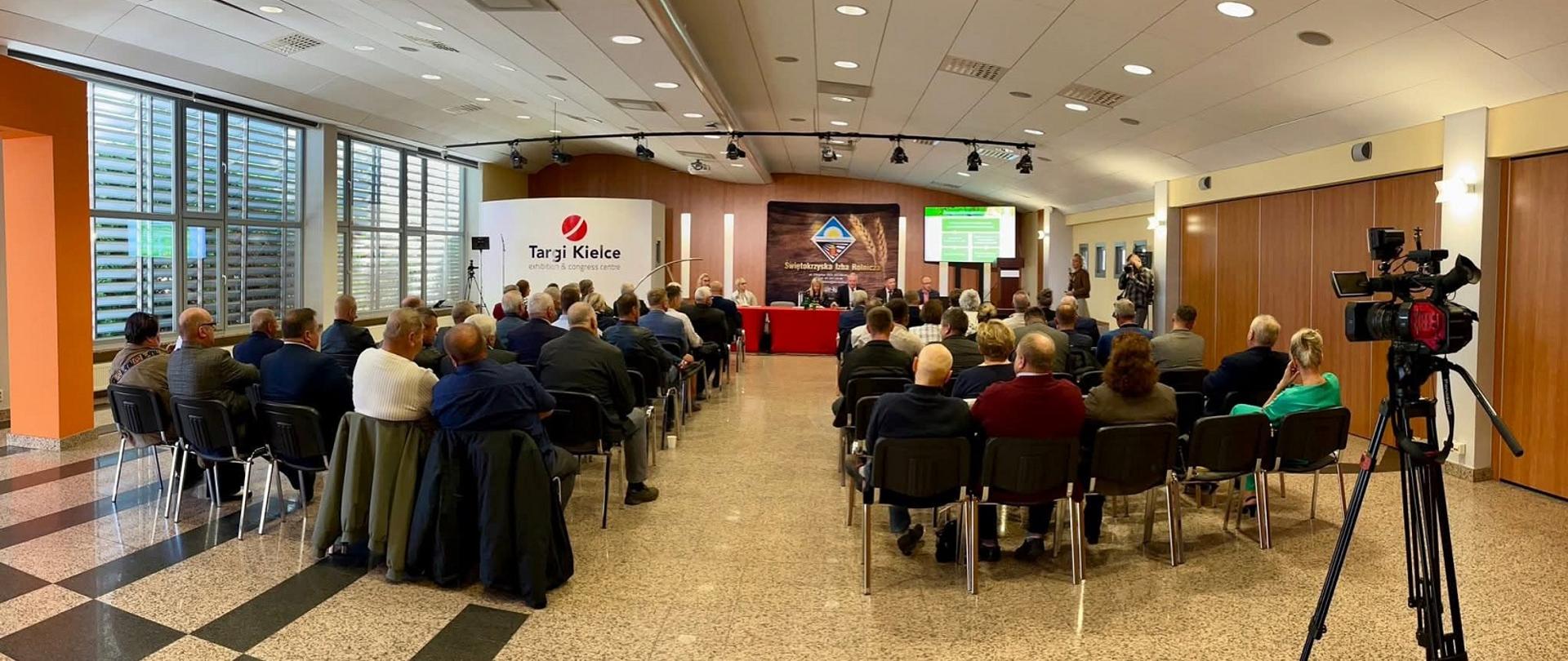 Zdjęcie przedstawia rzędy krzeseł na których siedzą uczestnicy Posiedzenia. W centralnej części fotografii roll-up z logo ŚIR oraz Targów Kielce. 