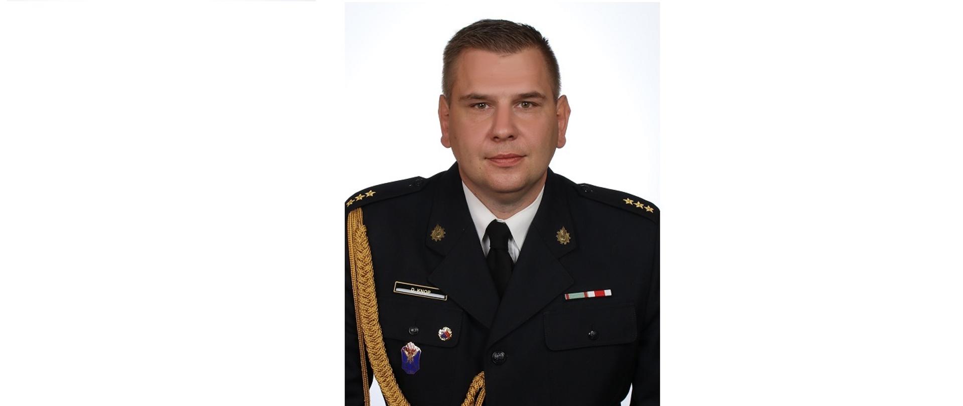 Na zdjęciu widzimy mężczyznę w wieku średnim. Ubrany jest w galowy czarny mundur strażacki, biała koszula, oraz czarny krawat. Na ramionach widzimy trzy złote gwiazdki, po prawej stronie zwisa złoty sznur. Mężczyzna uczesany jest na prawa stronę bez przedziałku. 